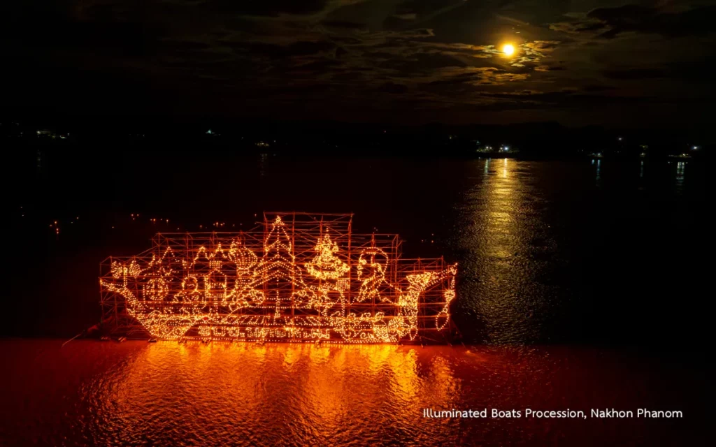 Un bateau traditionnel décoré de motifs religieux lumineux flotte sur le Mékong sous un ciel nocturne éclairé par la lune à Nakhon Phanom.