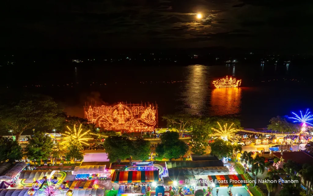 Vue panoramique nocturne des festivités du Lai Reua Fai avec des bateaux illuminés sur le Mékong, des attractions et des stands colorés sur les berges de Nakhon Phanom.