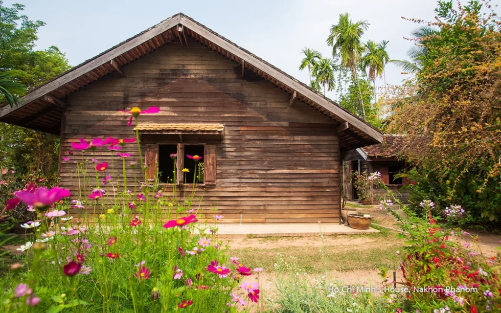 Traditioneel houten huis in de bloementuin van de historische locatie van Ho Chi Minh in Nakhon Phanom, met roze bloemen op de voorgrond en tropische bomen.