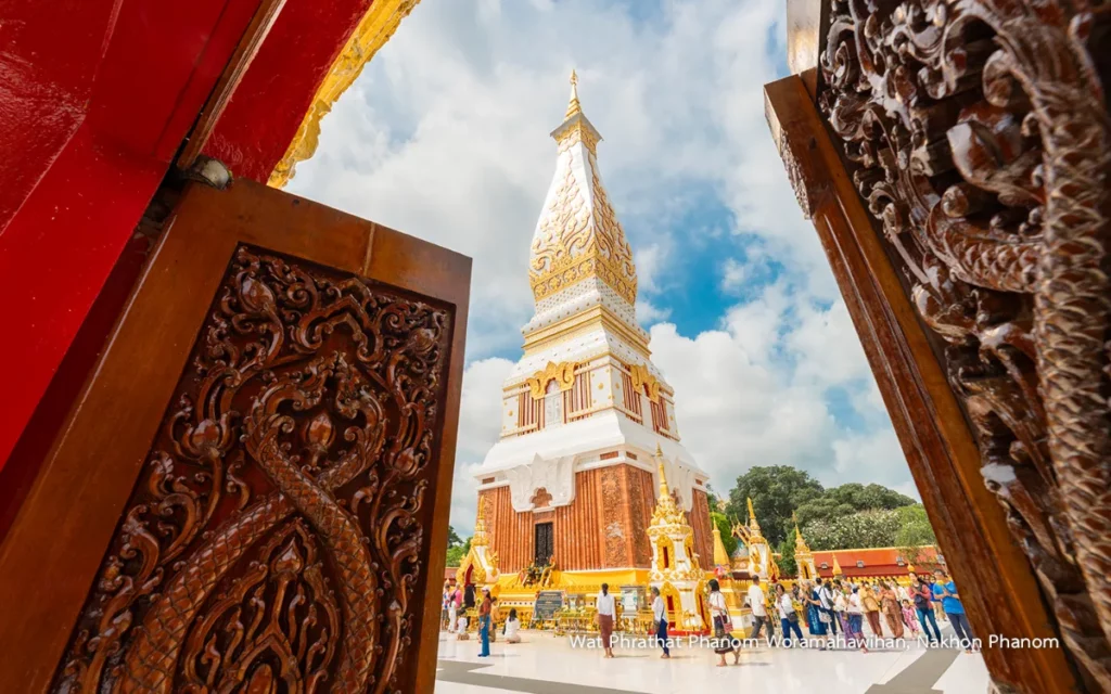 Majestueuze witte stoepa met gouden versieringen bij Wat Phra That Phanom, gefotografeerd door rijkelijk uitgesneden houten deuren, met bezoekers op het tempelterrein.