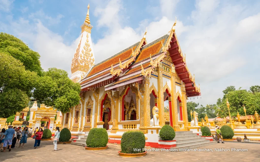 Bâtiment bouddhiste du Wat Phra That Phanom à Nakhon Phanom, avec toiture rouge et or, sculptures traditionnelles, colonnes ornées et grand stupa blanc en arrière-plan.
