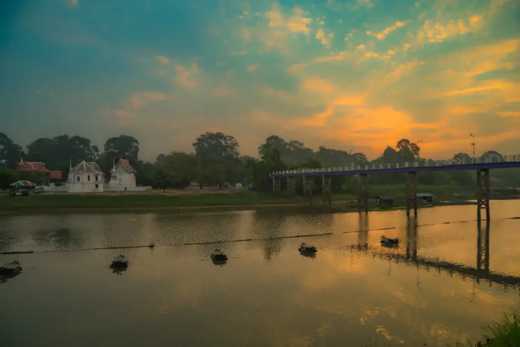 Een brug over de Sakae Krang-rivier bij zonsondergang, met witte tempelgebouwen aan de oever.