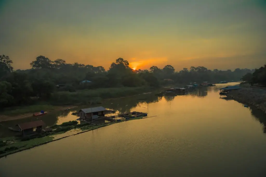 De Sakae Krang-rivier bij zonsondergang, met drijvende huizen op het water en bomen langs de oevers.