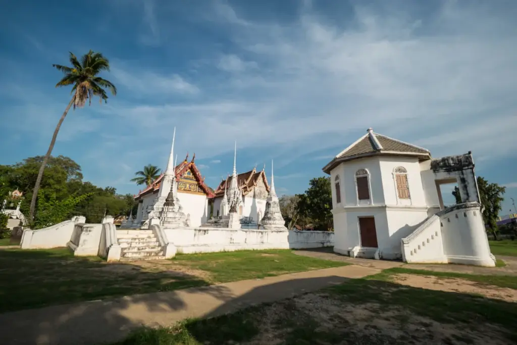 Witte gebouwen van Wat Uposatharam met verschillende chedi's, trappen en een palmboom onder de blauwe lucht.